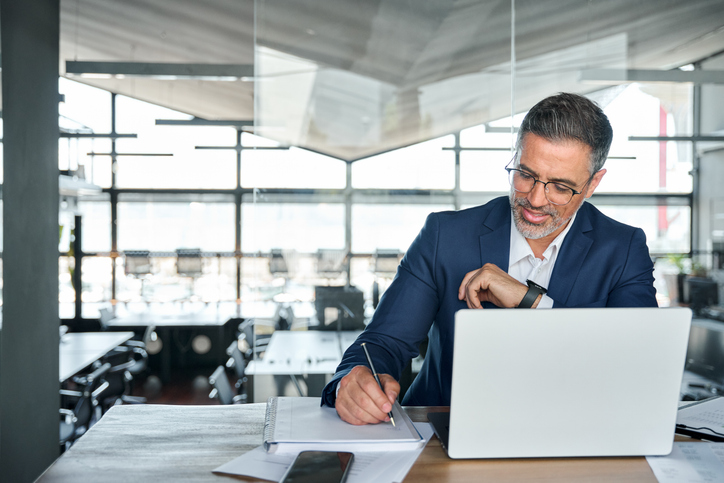 Middle aged smiling professional business man global company executive ceo manager or lawyer wearing suit sitting at desk in modern office working on laptop computer and writing notes, copy space.